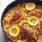 Plated one pot lemon herb chicken and rice with fresh parsley and lemon wedges on a wooden table.